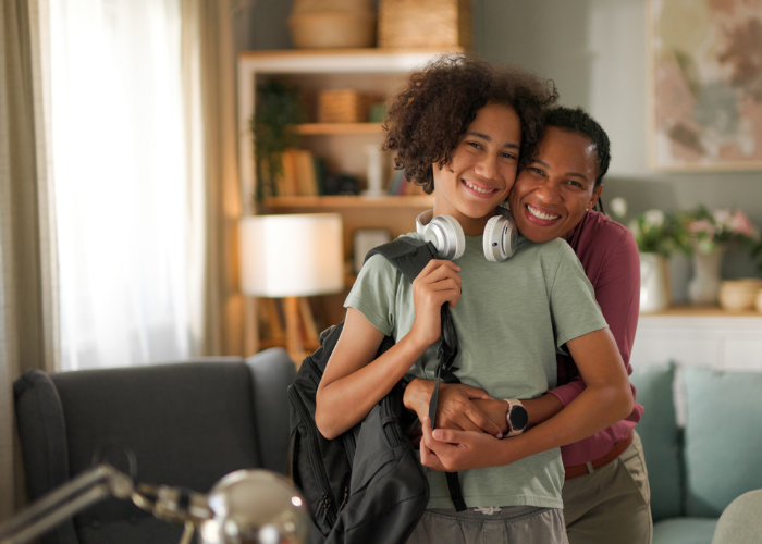 Joyful Reinvention with Maria Hendershot Smiling mother hugging her child who is wearing a backpack and headphones, ready for the first day of school at home.