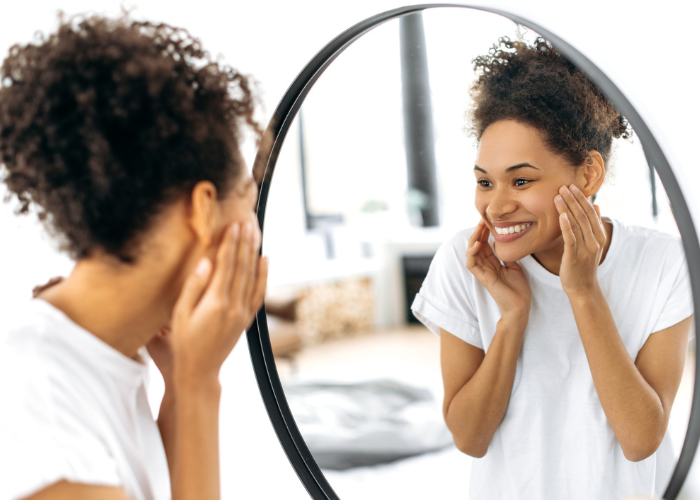 A woman smiling warmly at her reflection in a round mirror, symbolizing self-care, confidence, and valuing herself. Joyful Reinvention with Maria Hendershot