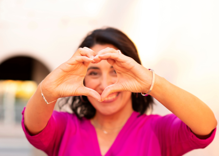 Woman smiling and forming a heart shape with her hands outdoors, wearing a bright pink top.