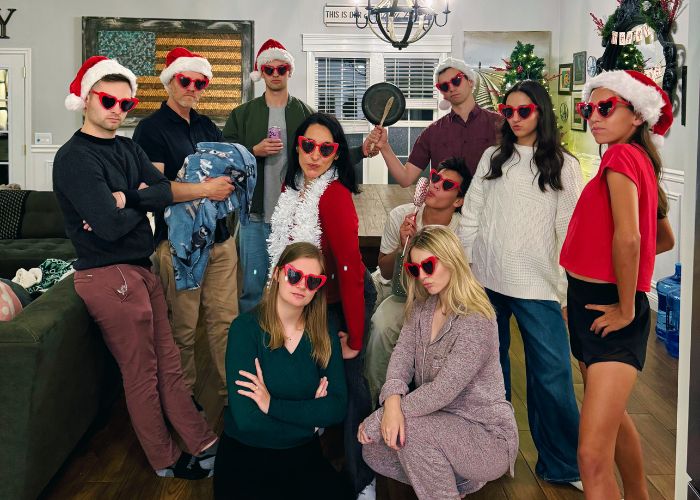 Family gathered wearing Santa hats and heart-shaped sunglasses posing playfully in a living room decorated for Christmas, during a family game and dance night.