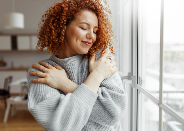 Woman with curly red hair gently hugging herself near a window, symbolizing self-love and emotional well-being.