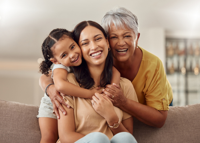 Three generations of women—a grandmother, mother, and young daughter—smiling and hugging together on a couch in a bright, cozy home.